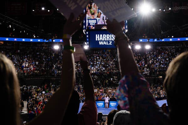 Kamala Harris stands onstage in an arena that is packed with supporters. The Jumbotron says “Harris-Walz.”