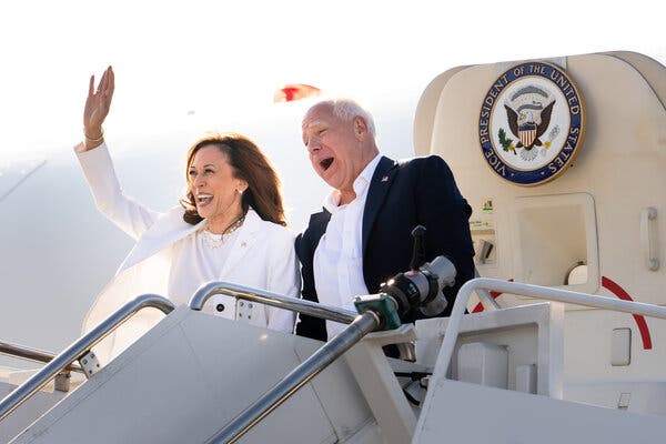 Kamala Harris waving and smiling with Tim Walz standing next to her, also smiling, as they walk out of Air Force Two in Detroit.