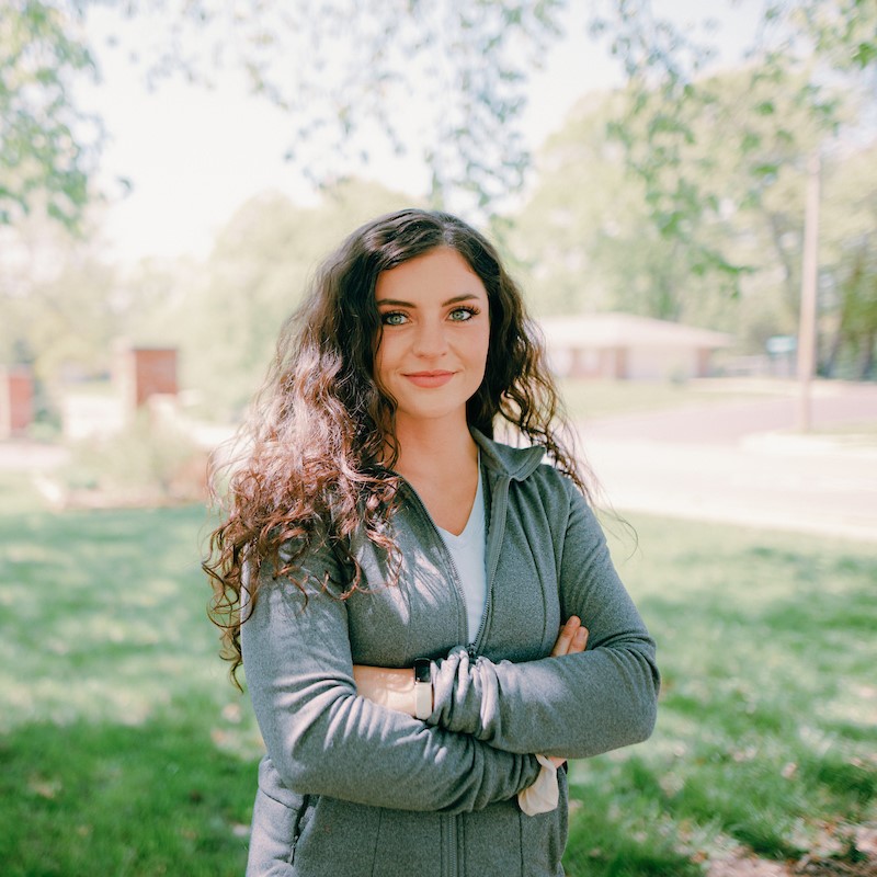 Undergraduate: Woman with long brown hair, standing outside, smiling at the camera with arms crossed.